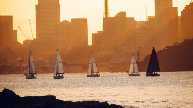Sunset photo with five yachts sailing in a line. Sydney skyscrapers are behind.