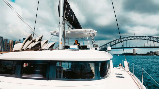 Looking back at the boat from the front. It has wrap-around windows and someone is steering it. The Sydney Harbour Bridge is behind.